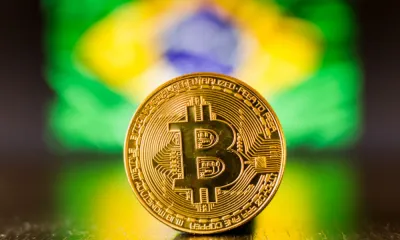 Foto de ua moeda de Bitcoin, e ao fundo, uma bandeira do Brasil desfocada