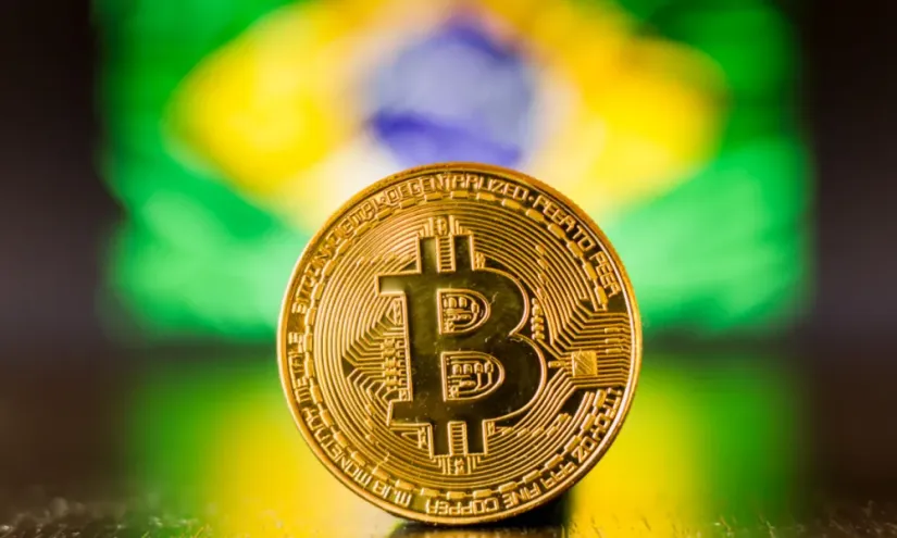 Foto de ua moeda de Bitcoin, e ao fundo, uma bandeira do Brasil desfocada