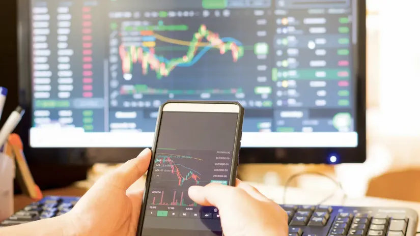 Pessoa negociando ativos digitais em um smartphone, com gráficos do mercado financeiro ao fundo.